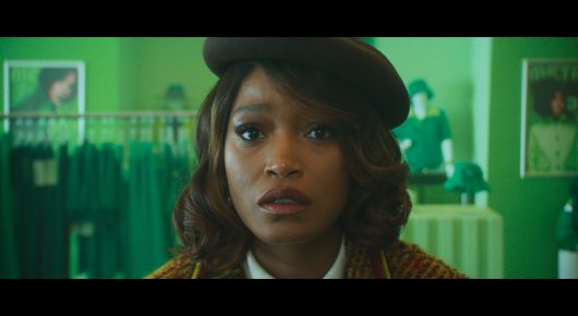 Still from I Love Boosters: a woman in a brown beret and tweed jacket looks worried, in a clothes shop lit green.