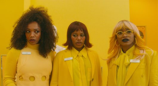 Still from I Love Boosters: three women in matching yellow jackets with name badges stand side by side against a yellow wall.