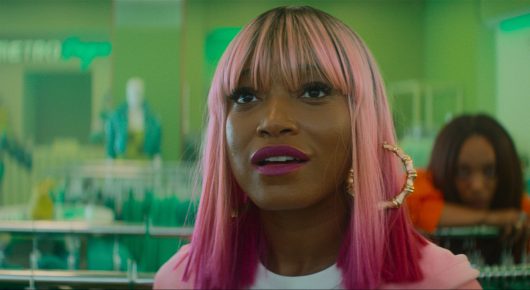 Still from I Love Boosters: a woman with pink hair and a fringe looks up and to one side, in a shop with green racks of clothes, while another woman rests her head on her arm behind her.