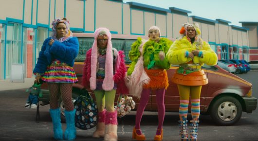 Still from I Love Boosters: four women in brightly coloured fluffy coats and patterned tights stand in front of an old red car in a car park.