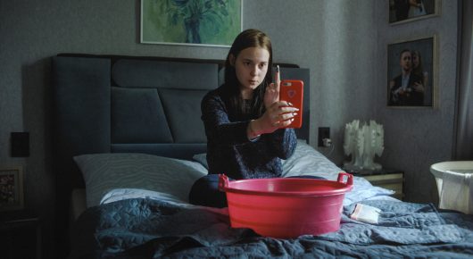 Still from I Rarely Wake Up Dreaming: a young woman sits on a bed holding a red item up in front of her, with a pink washing-up bowl before her, in a grey bedroom.