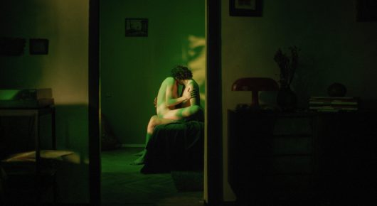 Still from I Rarely Wake Up Dreaming: a person sits hunched in a green-lit doorway of a dim apartment.