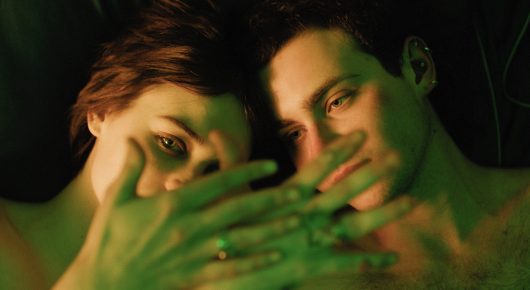 Still from I Rarely Wake Up Dreaming: two young men press their faces together with a hand held across their mouths, in green light.