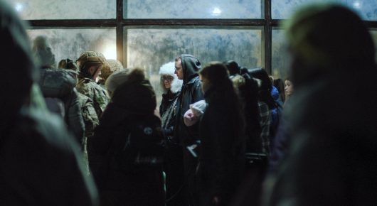 Still from I Rarely Wake Up Dreaming: a crowd of people in dark winter clothing stands in a dim room in front of large windows.