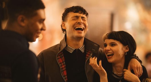 Still from I See Buildings Fall Like Lightning: a young man laughs with his eyes closed beside a young woman with a dark bob who is also laughing, with another man in profile at the left.