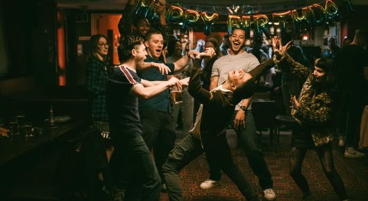 Still from I See Buildings Fall Like Lightning: people dance in a crowded bar decorated with balloons and banners.