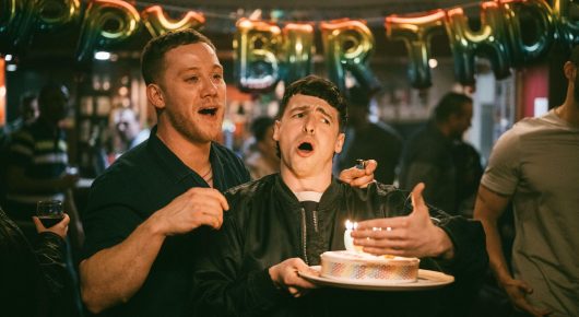 Still from I See Buildings Fall Like Lightning: a man looks astonished in front of a lit birthday cake held up by another man, in a bar with a birthday banner.