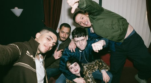 Still from I See Buildings Fall Like Lightning: a group of young people squeeze into a selfie, some leaning over each other, in a dim room.