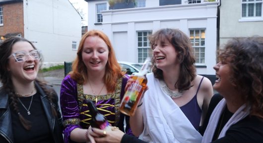 Still from Cambridge Connections: four young people laugh together on a street, one holding a bottle and one in a white toga-style costume.