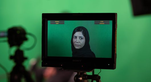 Still from It Means Hope: a monitor in front of a green screen shows a woman in a headscarf, with a camera beside it.