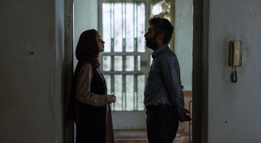 Still from It Means Hope: a woman in a headscarf and a man stand facing each other in a narrow, dim hallway beside a barred window.