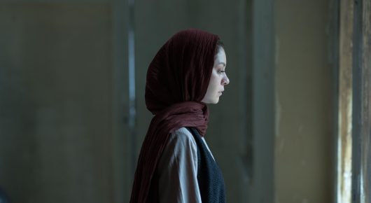 Still from It Means Hope: a woman in a red headscarf seen in profile in a dim interior.
