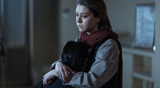 Still from It Means Hope: a woman in a red scarf holds a child close to her and looks down with a serious expression.