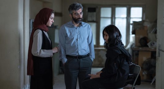 Still from It Means Hope: three people stand and sit together in a bright room full of boxes, one of them wearing a red headscarf.
