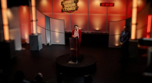 Still from Jasmine. Home. Mother.: a wide view of an orange and red game show set, with a woman in red at a podium and a man standing to the side.