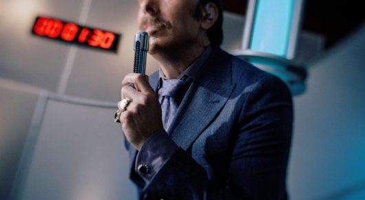 Still from Jasmine. Home. Mother.: a man in a blue suit holds up a small black object near his face on a brightly lit game show set, with a digital countdown clock behind him.