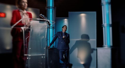 Still from Jasmine. Home. Mother.: two people stand on a game show stage with glass podiums and a blue-lit set, one in a red outfit and one in a dark suit.