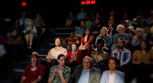 Still from Jasmine. Home. Mother.: a studio audience of mixed ages sits in tiered seating under a digital clock.