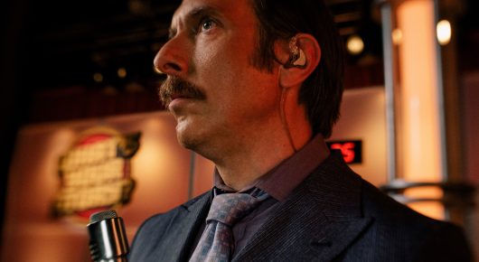 Still from Jasmine. Home. Mother.: a man in a grey suit and tie with an earpiece looks up and to one side, with a microphone in front of him.