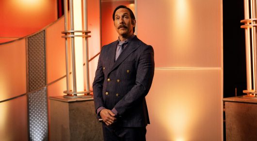 Still from Jasmine. Home. Mother.: a man in a navy double-breasted suit stands in front of an orange and cream panelled game show set.