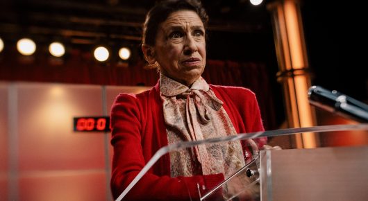 Still from Jasmine. Home. Mother.: a woman in a red jacket and ruffled cream blouse stands at a clear podium under bright stage lights, looking to one side with a tense expression.