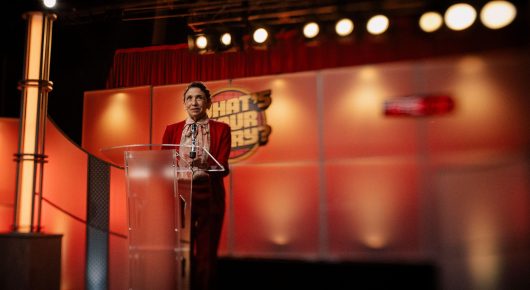 Still from Jasmine. Home. Mother.: a woman in red stands at a clear podium on a stage with an orange backdrop and a row of lights above.