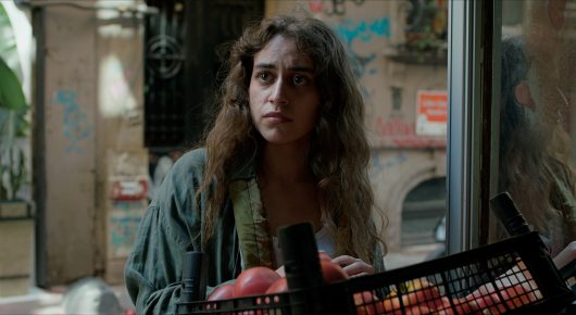 Still from Krizalit: a young woman with wavy brown hair stands by a window holding a black basket, with a city street behind her.