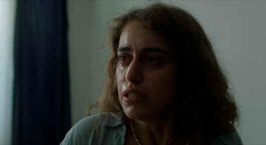 Still from Krizalit: a close-up of a young woman with wavy brown hair looking to one side with a worried expression.