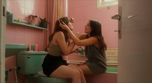 Still from Krizalit: two young women sit on the edge of a bath in a pink-tiled bathroom, one touching the other's face.