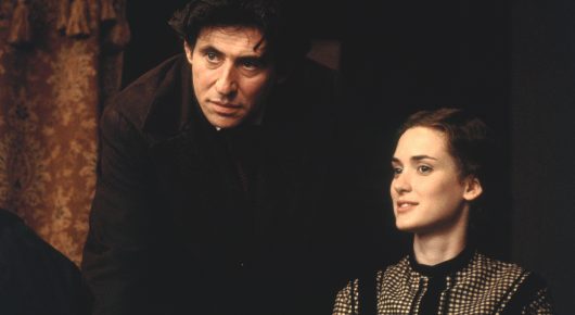 Still from Little Women: a man in dark clothes and a young woman in a dark patterned dress sit side by side in a dim room.