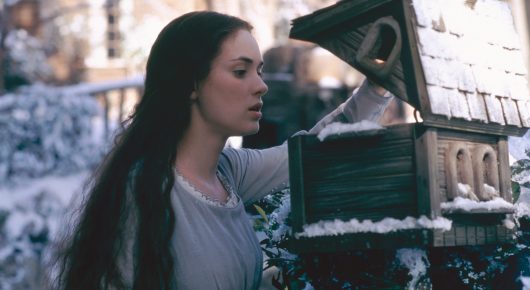 Still from Little Women: a young woman with long dark hair stands in the snow beside a small wooden post box.
