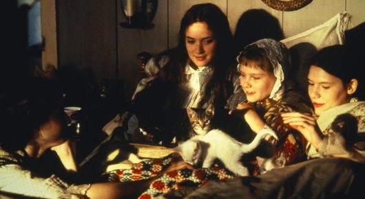 Still from Little Women: several girls in nightclothes and headscarves lie in bed, one with a white cat, under a patchwork quilt.