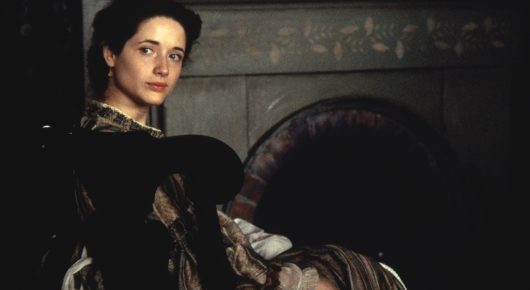 Still from Little Women: a young woman stands in a dim room beside a large stone fireplace.
