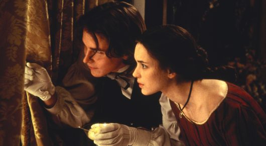 Still from Little Women: a young man and a woman in 19th-century dress lean in to look at something he holds in his gloved hand, beside heavy curtains.