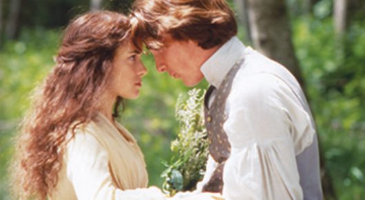 Still from Little Women: a young woman with long wavy hair and a man in a waistcoat touch foreheads in a sunlit garden.