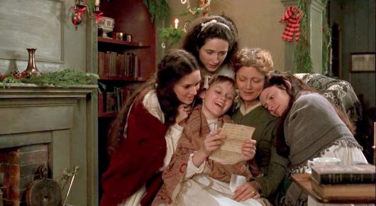 Still from Little Women: a group of women and girls in 19th-century dress gather close around a letter beside a fireplace decorated with Christmas garlands.