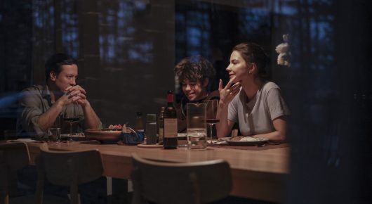 Still from Minotaur: four people sit around a dinner table in a dimly lit room, seen from outside the window.