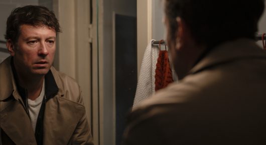 Still from Minotaur: a man in a brown jacket looks off to one side in a dim kitchen, with another person's shoulder in the foreground.