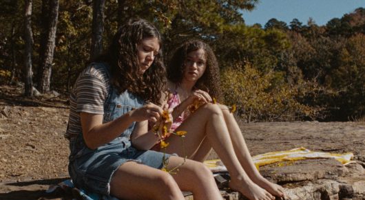 Still from Mouse: two girls with curly hair sit on rocks in a wood, one in a denim dress, both looking down at something in their hands.