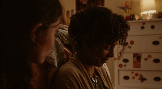 Still from Mouse: a close-up of a girl with curly hair looking down, beside a chest of drawers with stickers, in warm lamp light.