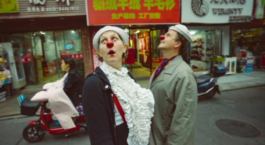 Still from My Family and Other Clowns: two people in clown costume, one with a red nose and a white ruffled shirt and the other in a hat and green coat, stand looking up on a busy street with shop signs behind them.