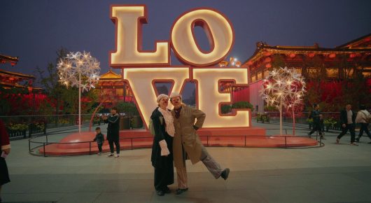Still from My Family and Other Clowns: two people in long coats stand in front of a large illuminated sign reading LOVE in a plaza at dusk.
