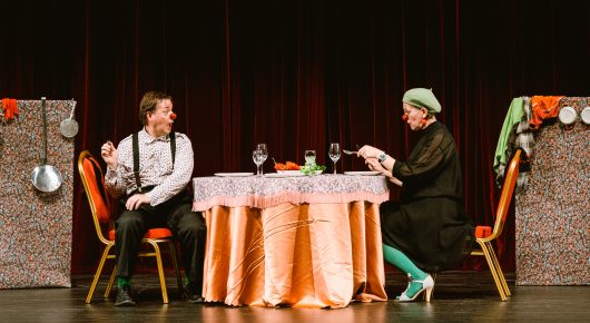 Still from My Family and Other Clowns: two performers in costume sit at a round table with an orange cloth and wine glasses on a theatre stage with red curtains.