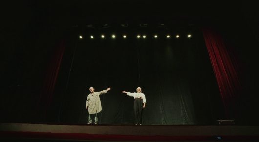 Still from My Family and Other Clowns: two performers in light clothes stand far apart on a dark stage with spotlights above and red curtains at the sides.