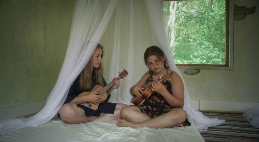 Still from My Family and Other Clowns: two women sit on a bed under a white mosquito net, each playing a small stringed instrument, beside a window.