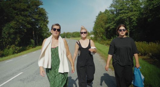 Still from My Family and Other Clowns: three women in sunglasses walk along an empty road lined with trees.