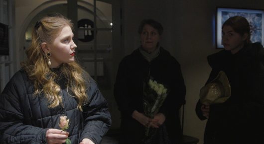 Still from My Family and Other Clowns: a young woman with long wavy hair and a dark quilted coat looks to one side in a dim corridor, with two other people behind her, one holding flowers.