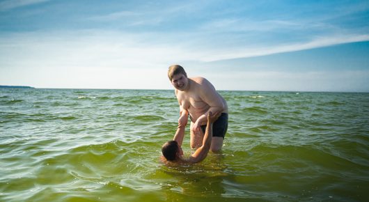 Still from My Family and Other Clowns: a man stands knee-deep in a lake, bending forward over a dark bucket.