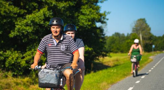 Still from My Family and Other Clowns: a man in a striped polo shirt and a helmet cycles along a country road, with another person behind him and a cyclist further back.