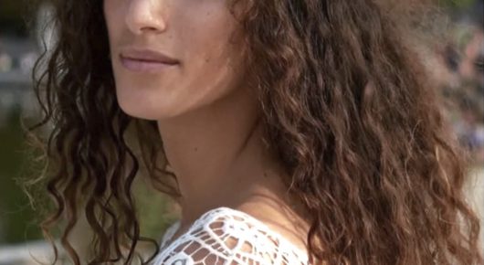 Still from My Father and Qaddafi: a close-up of a woman with long curly hair, her face cropped at the nose, wearing a white lace top.
